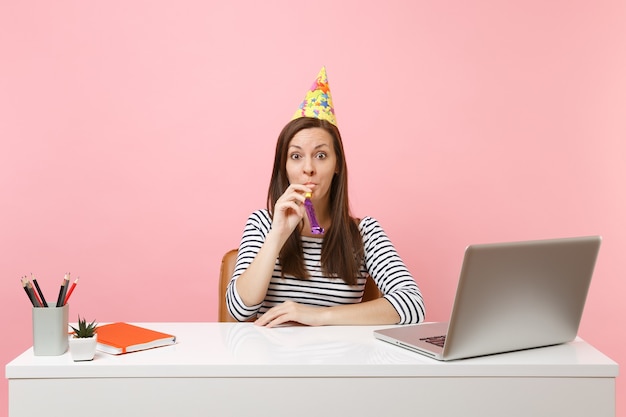Joven sorprendida en sombrero de fiesta de cumpleaños con pipa jugando celebrando mientras se sienta a trabajar en el escritorio blanco con computadora portátil pc aislada sobre fondo rosa pastel. Concepto de carrera empresarial de logro. Copie el espacio.