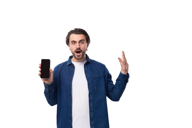 Foto joven sorprendido apuesto moreno brutal tipo caucásico muestra pantalla de teléfono inteligente con maqueta