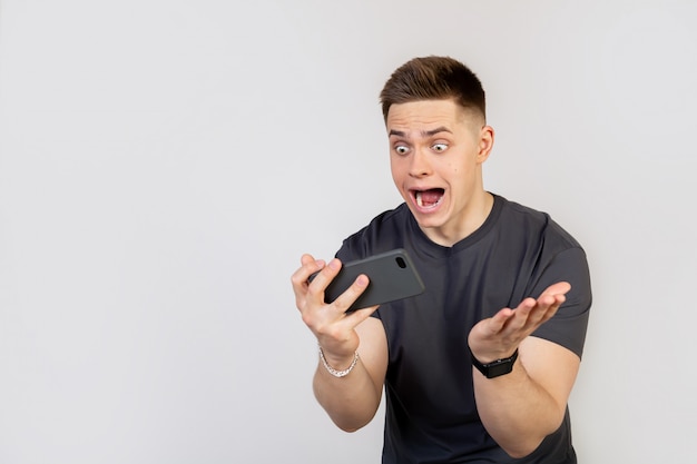 Un joven sorprendido y conmocionado sostiene un teléfono inteligente en sus manos y mira la pantalla con la boca abierta y los ojos grandes