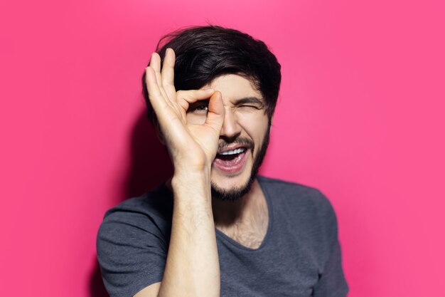 joven sorprendido, mirando a través del gesto ok con la mano, aislado en la pared de color rosa coral.