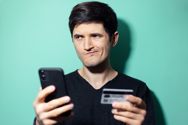 Joven sorprendido con smartphone y tarjeta de crédito en la mano en la pared de color aqua menthe.