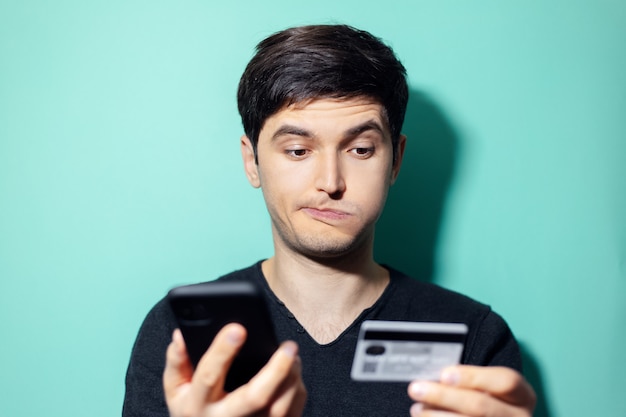 joven sorprendido con smartphone y tarjeta de crédito en la mano en la pared de color aqua menthe.