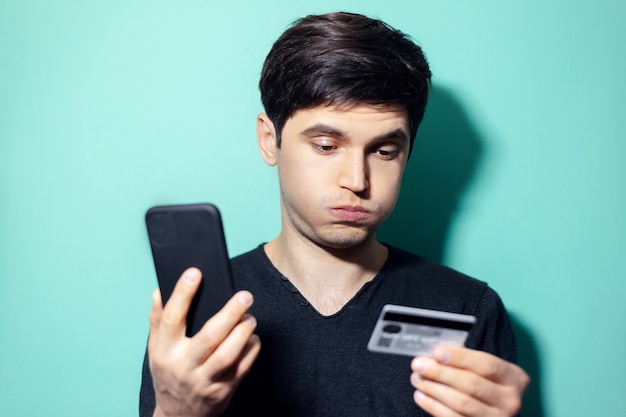 joven sorprendido con smartphone y tarjeta de crédito en la mano en la pared de color aqua menthe.