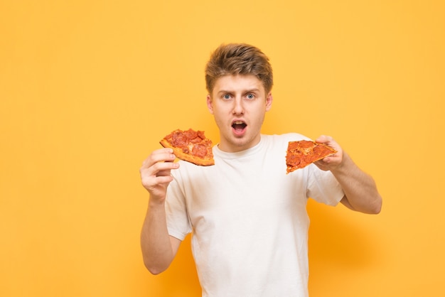 Joven sorprendido sobre un fondo amarillo, sosteniendo dos piezas de pizza en sus manos