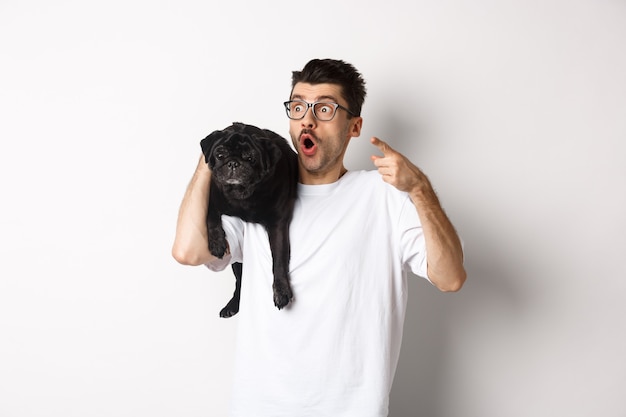 Joven sorprendido sosteniendo un lindo perro negro en el hombro, señalando con el dedo a la izquierda en la oferta promocional, mirando impresionado y sin palabras, de pie sobre blanco.