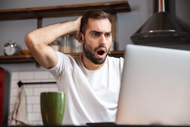 Joven sorprendido usando la computadora portátil mientras está sentado en la mesa de la cocina