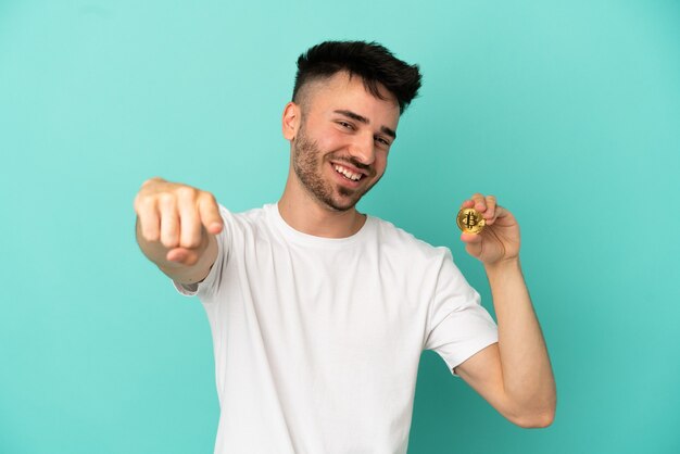 Joven sosteniendo un Bitcoin aislado sobre fondo azul apuntando al frente con expresión feliz