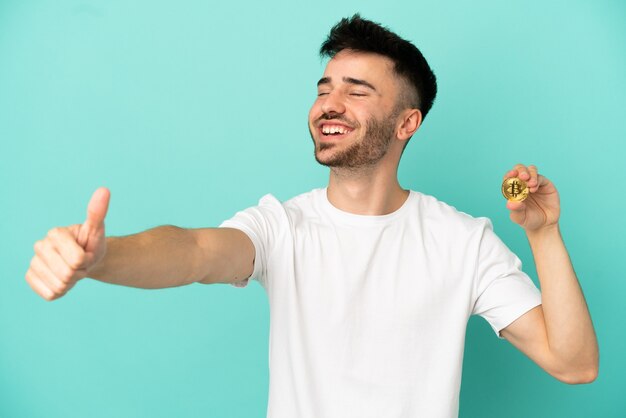 Joven sosteniendo un Bitcoin aislado sobre fondo azul dando un gesto de pulgar hacia arriba