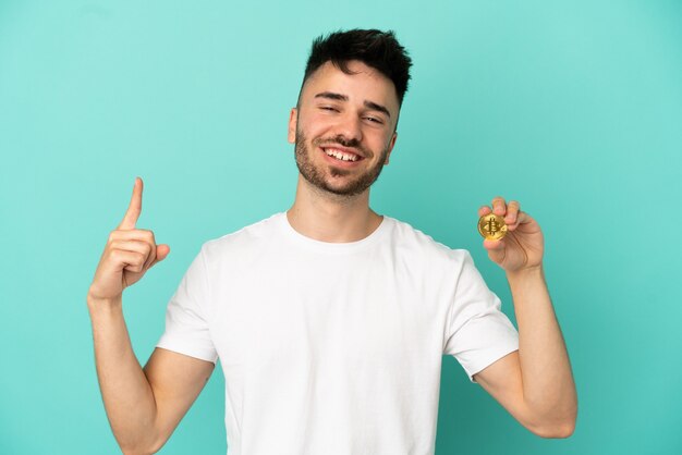 Joven sosteniendo un Bitcoin aislado sobre fondo azul mostrando y levantando un dedo en señal de lo mejor