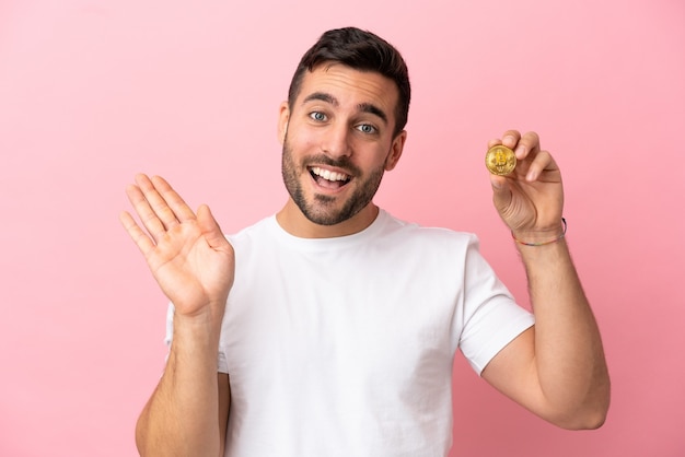 Joven sosteniendo un Bitcoin aislado sobre fondo rosa saludando con la mano con expresión feliz