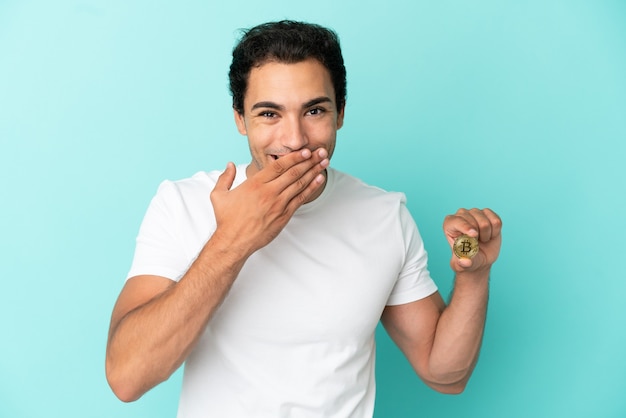 Joven sosteniendo un Bitcoin sobre fondo azul aislado feliz y sonriente cubriendo la boca con la mano