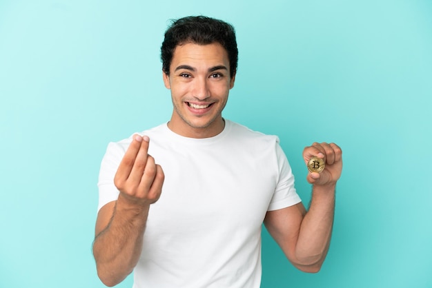 Joven sosteniendo un Bitcoin sobre fondo azul aislado haciendo gesto de dinero