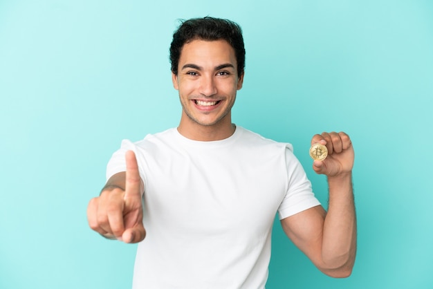 Joven sosteniendo un Bitcoin sobre fondo azul aislado mostrando y levantando un dedo