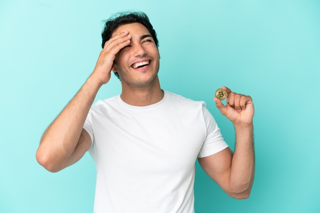 Joven sosteniendo un Bitcoin sobre fondo azul aislado sonriendo mucho