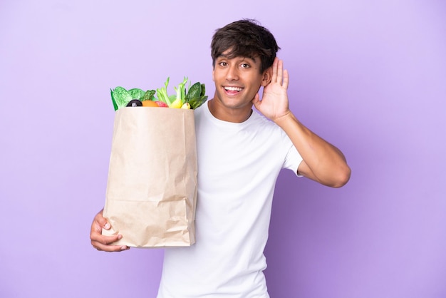 Joven sosteniendo una bolsa de compras aislada en un fondo morado escuchando algo poniendo la mano en la oreja