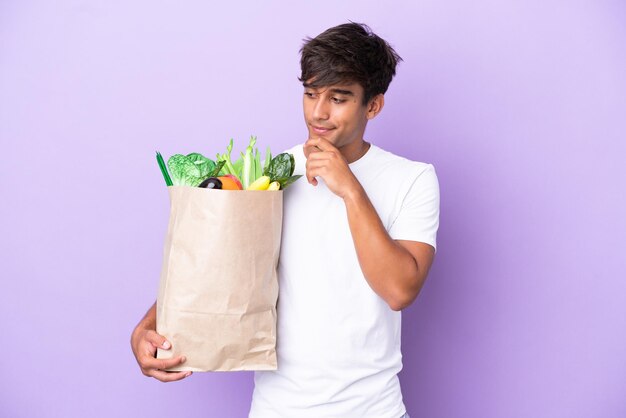 Joven sosteniendo una bolsa de compras aislada en un fondo morado mirando hacia un lado y sonriendo