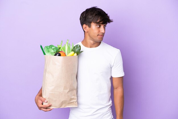 Joven sosteniendo una bolsa de compras aislada en un fondo morado mirando hacia un lado