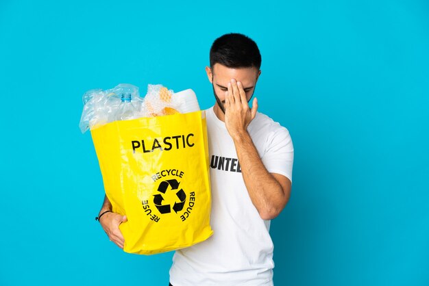 Joven sosteniendo una bolsa llena de botellas de plástico para reciclar aislado en la pared azul con expresión cansada y enferma