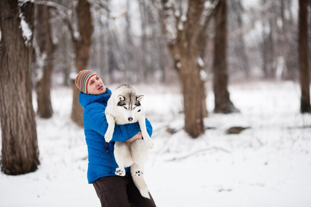 Joven sosteniendo un cachorro de husky en el bosque de invierno. Disparo horizontal al aire libre.