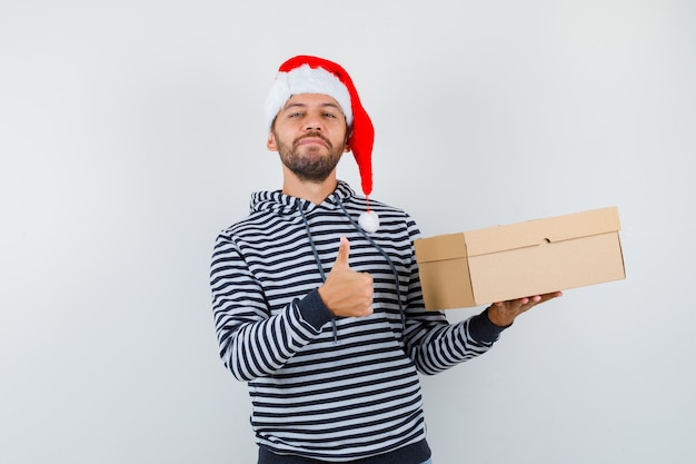 Joven sosteniendo una caja de cartón, mostrando el pulgar hacia arriba en una sudadera con capucha, un sombrero de Santa Claus y luciendo confiado. vista frontal.