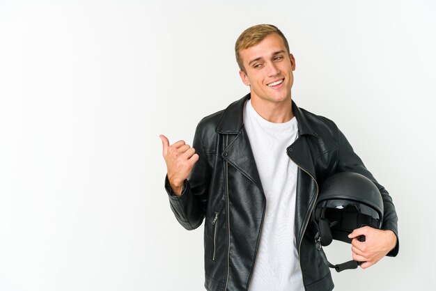 Joven sosteniendo un casco de moto aislado en puntos de pared blanca con el dedo pulgar lejos, riendo y despreocupado