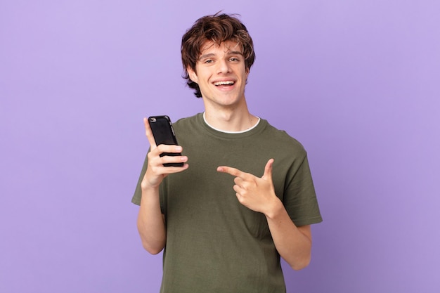 Joven sosteniendo una celda sonriendo alegremente, sintiéndose feliz y apuntando hacia un lado