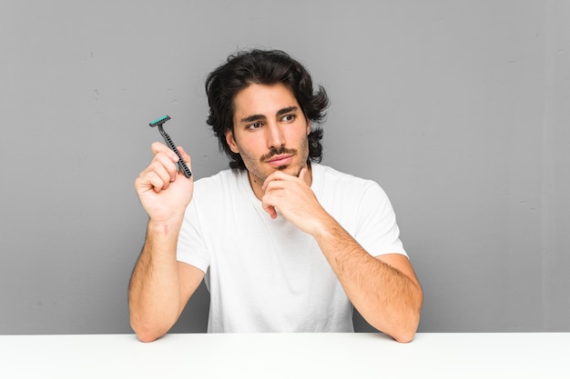 Joven sosteniendo una hoja de afeitar mirando hacia los lados con expresión dudosa y escéptica.