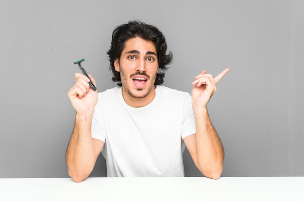 Joven sosteniendo una hoja de afeitar sonriendo alegremente señalando con el dedo de distancia.