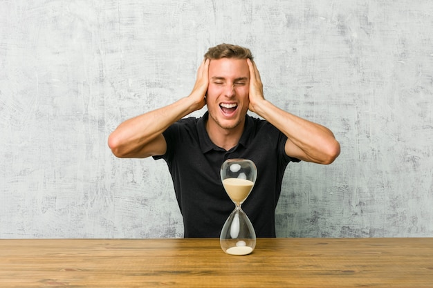 Foto joven sosteniendo un reloj de arena en una mesa ríe alegremente manteniendo las manos sobre la cabeza. concepto de felicidad