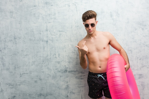 Joven sosteniendo una rosquilla inflable rosa con un traje de baño apuntando con el dedo hacia ti como invitando a acercarse.