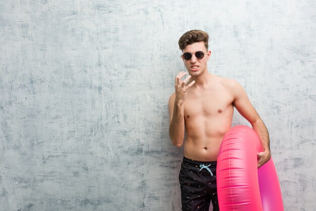 Joven sosteniendo una rosquilla inflable rosa vistiendo un traje de baño molesto gritando con las manos tensas.