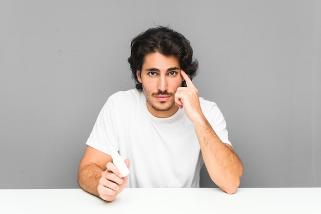 Joven sosteniendo un spray nasal apuntando su sien con el dedo, pensando, centrado en una tarea.