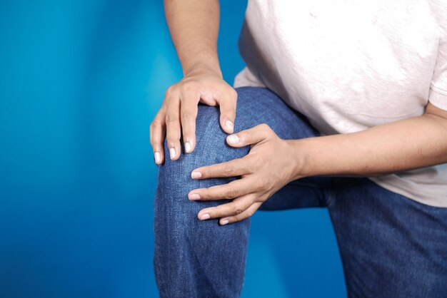 Joven sosteniendo su rodilla por sufrir dolor en las articulaciones contra el fondo azul.