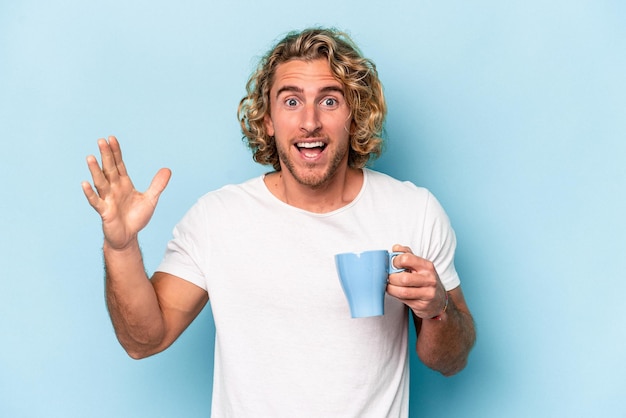 Joven sosteniendo una taza aislada de fondo azul recibiendo una agradable sorpresa, emocionado y levantando la mano.