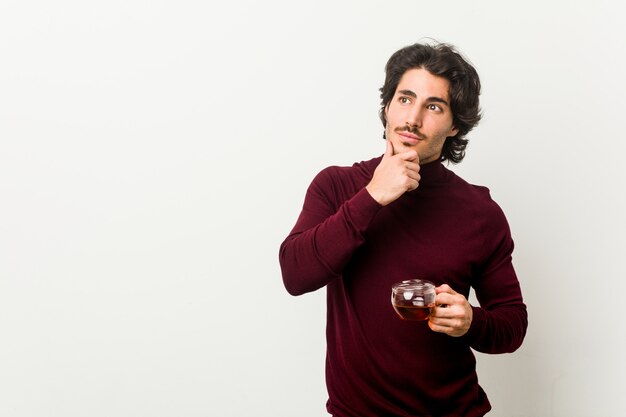 Joven sosteniendo una taza de té mirando hacia los lados con expresión dudosa y escéptica.