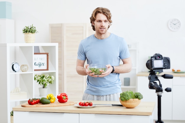 Joven sosteniendo un tazón con ensalada y diciendo cómo cocinar ensalada de verduras en línea en la cámara en la cocina