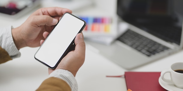 Joven sosteniendo el teléfono inteligente de pantalla en blanco en un lugar de trabajo cómodo con papelería