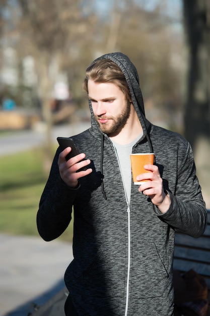 El joven sostiene una taza de papel de café o té y un teléfono móvil o celular en una camiseta con capucha soleada al aire libre en el parque sobre un fondo natural borroso blogueando por la energía de la mañana y navegando por Internet