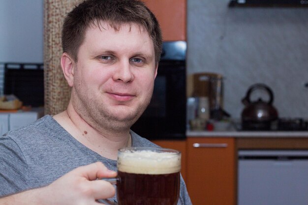 Un joven sostiene un vaso de cerveza oscura sentado en su cocina.