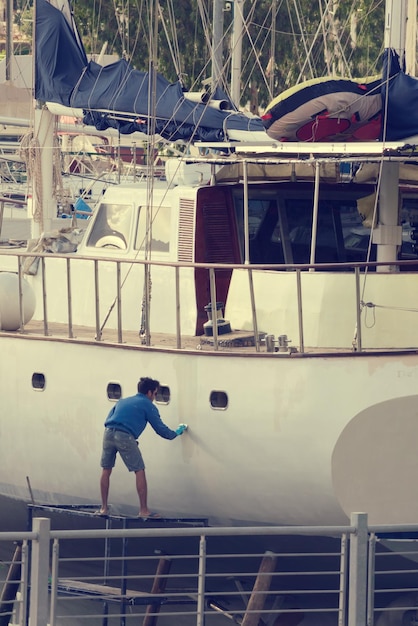 Un joven con suéter azul y pantalones cortos pinta el casco del barco