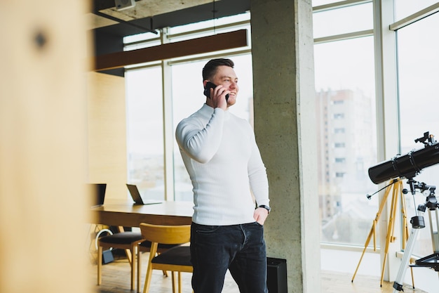 Un joven con un suéter blanco está parado en una oficina espaciosa y hablando por teléfono Un hombre habla con sus colegas a través de un enlace de video