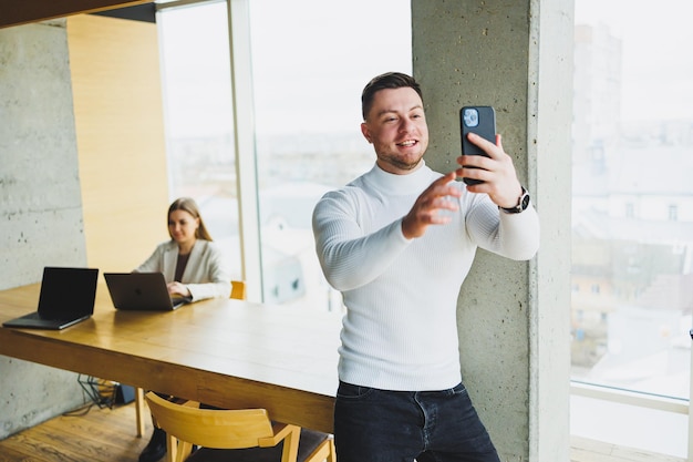 Un joven con un suéter blanco está parado en una oficina espaciosa y hablando por teléfono Un hombre habla con sus colegas a través de un enlace de video