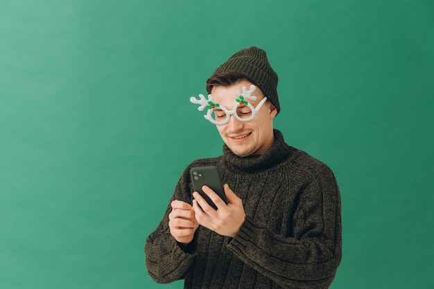 Un joven con un suéter cálido, gafas de carnaval y un teléfono en las manos aislado en un fondo verde