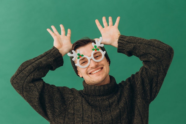 Un joven con un suéter cálido, una gorra y gafas de carnaval aisladas en un fondo verde