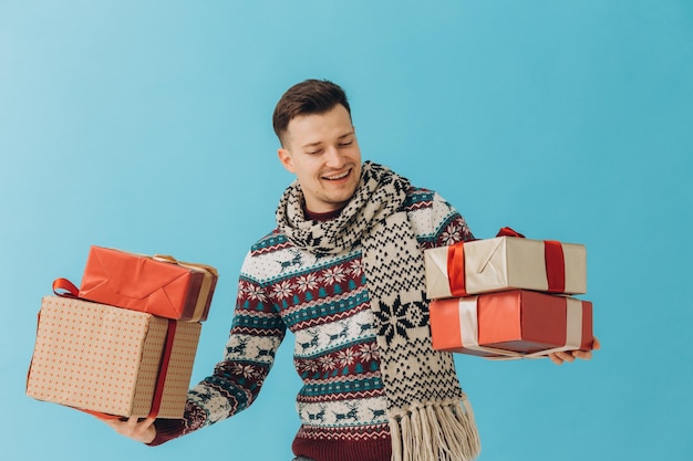 Joven en suéter de Navidad y bufanda sosteniendo muchas cajas de regalo con lazo de cinta de regalo aislado sobre fondo azul Concepto de celebración de feliz año nuevo