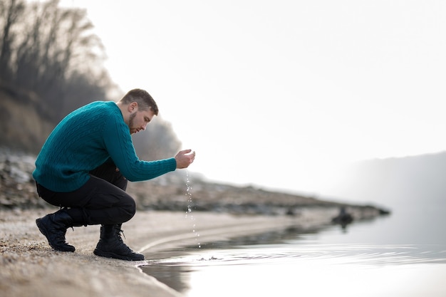 Un joven con un suéter turquesa se moja las manos en el río.