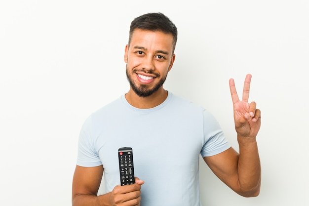 Joven del sur de Asia sosteniendo un controlador de tv alegre y despreocupado mostrando un símbolo de paz con los dedos.