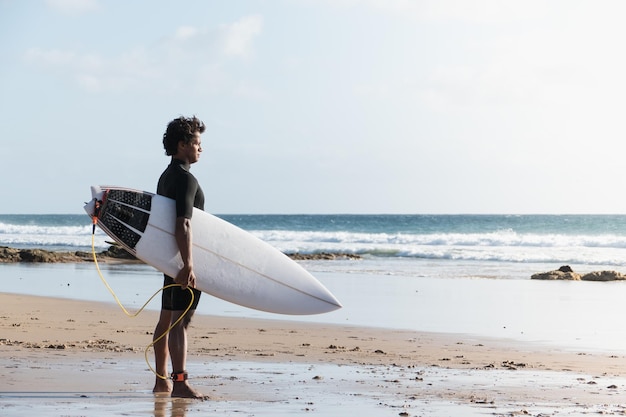 Joven surfista africano parado en la orilla de la playa mirando las olas