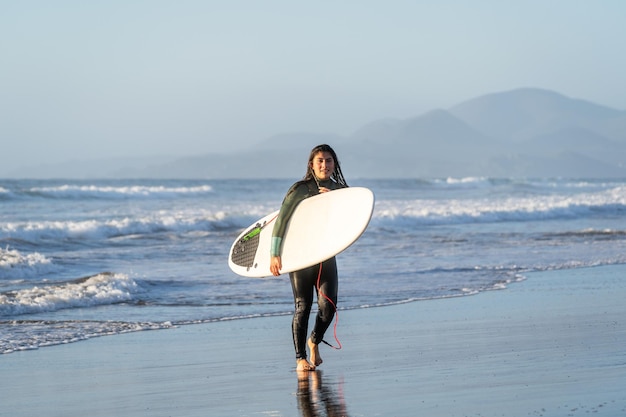 Joven surfista latino caminando por la playa con tabla de surf