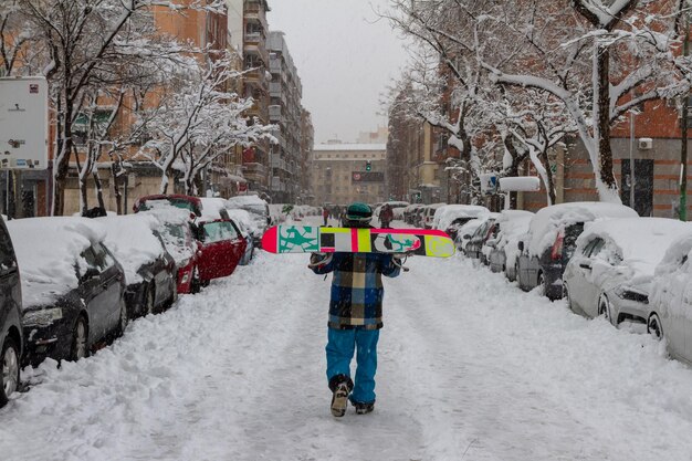 un joven con una tabla de snowboard camina por una ciudad nevada